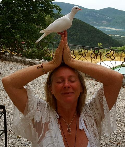 A brown-haired old woman standing yoga pose and a Pigeon is sitting at her head, her house surrounded by a natural mountain area